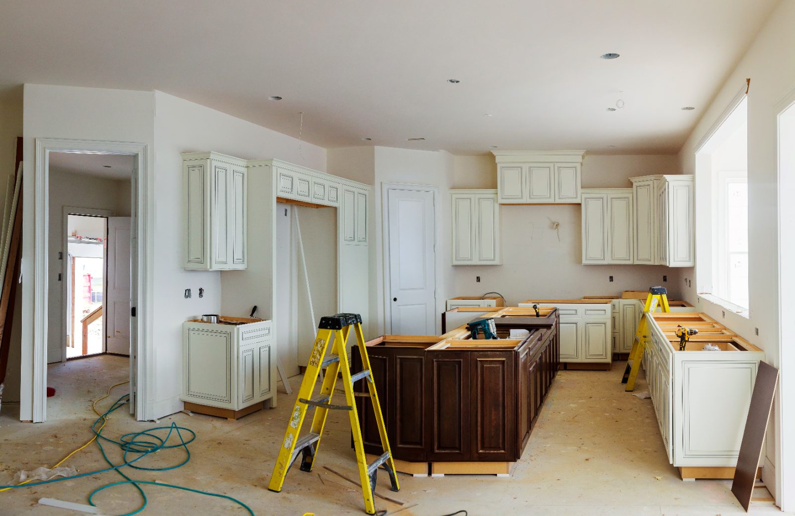 Kitchen under construction with cream cabinets, dark island, yellow ladders, and tools